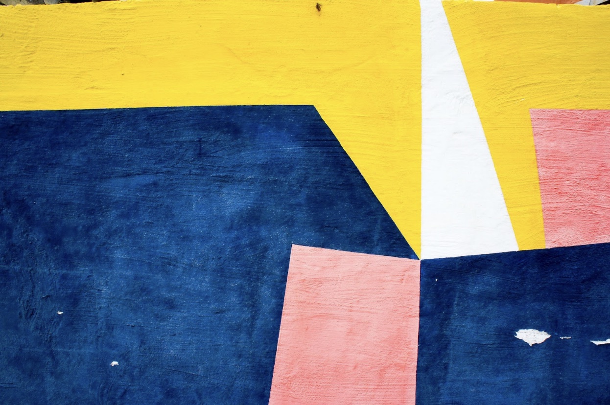 Seven painted abstract angular yellow pink and blue shapes of varying size, converging to the right of the middle of the canvas